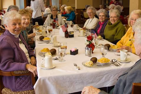 A group of elderly women sitting around a long dining table in a senior living facility, smiling and enjoying a meal together. The table is set with plates of food, glasses of iced tea, coffee cups, and small American flags as decorations.