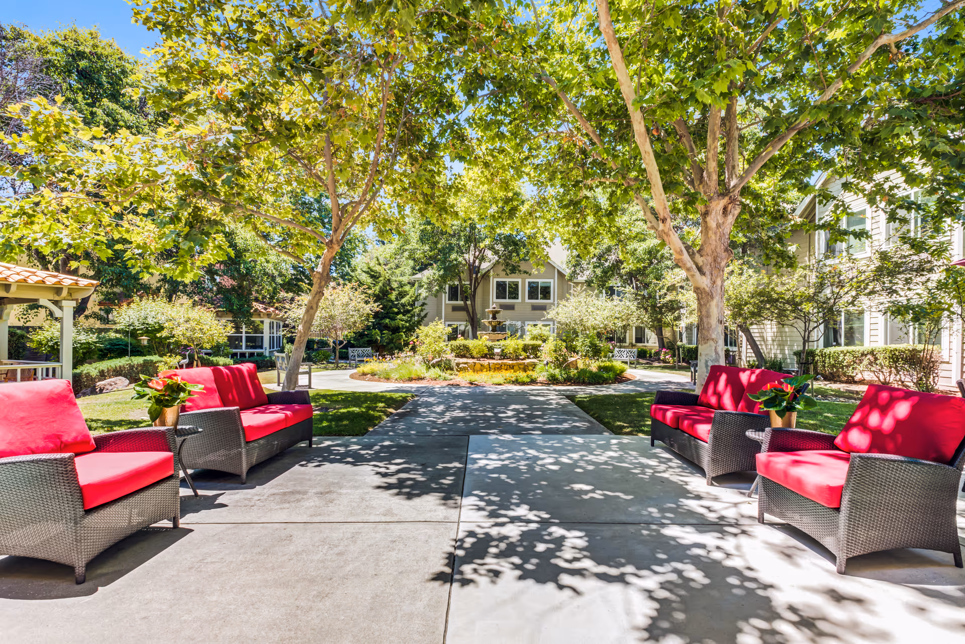 Outdoor seating area with four wicker armchairs featuring bright red cushions arranged on a concrete patio under large leafy trees. In the background, there is a circular garden with a fountain surrounded by greenery and residential buildings.