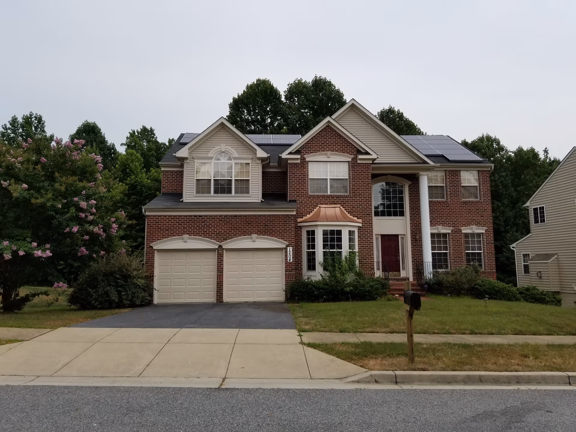 Front exterior view of a two-story brick house with a double garage, a red front door, white columns, and solar panels on the roof. The house is surrounded by greenery and trees, with a mailbox on the lawn near the sidewalk.