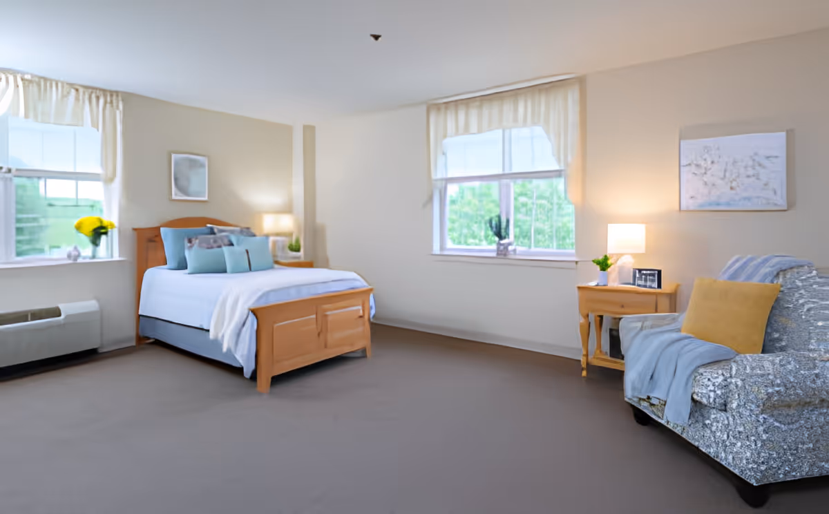 Bright furnished bedroom with a wooden bed, armchair, nightstands, and two windows.