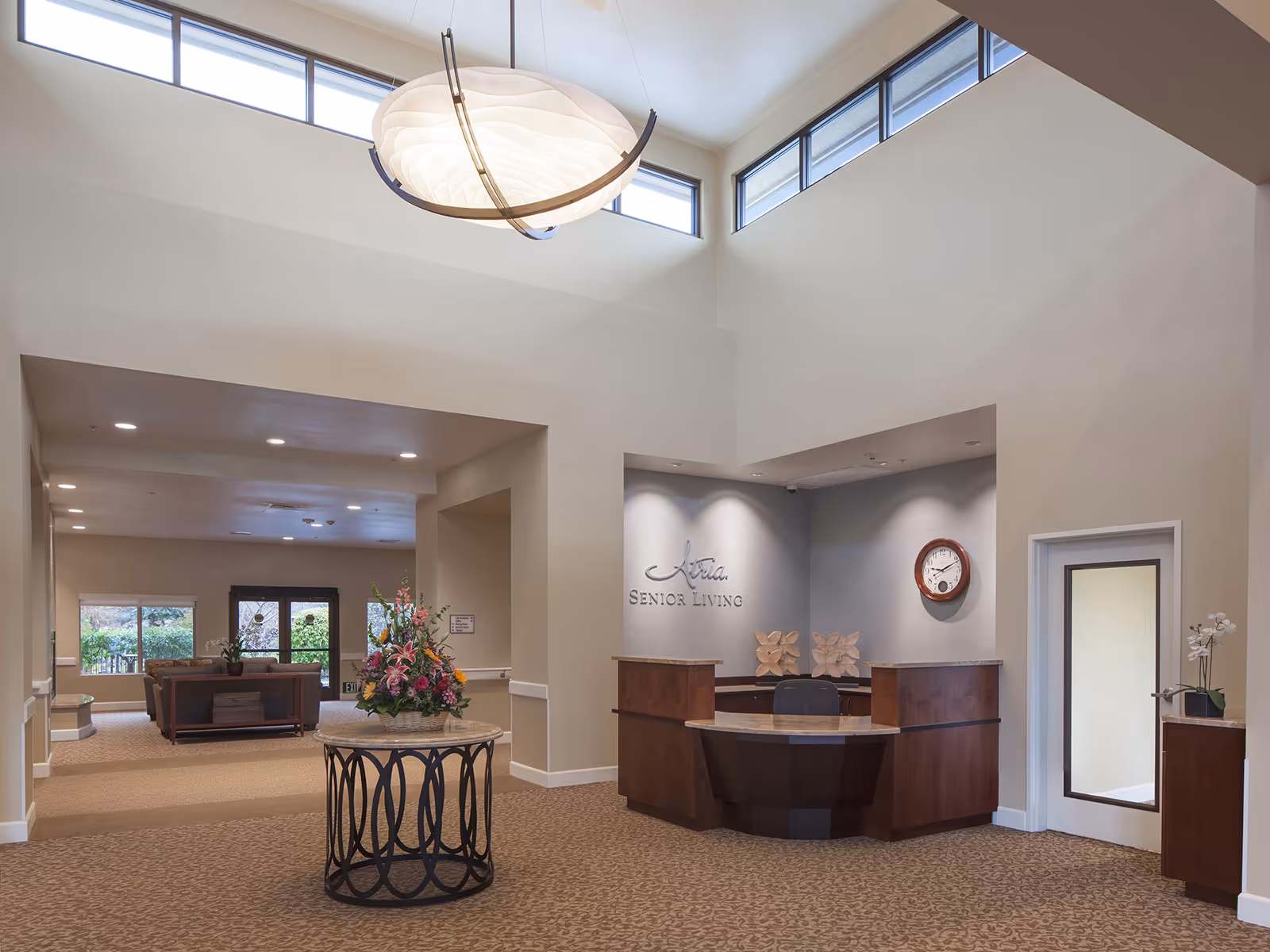 Reception area of Atria Walnut Creek senior living facility with a wooden front desk, a round table with a floral arrangement in the center, beige carpet, high ceilings with windows near the ceiling, and a seating area visible in the background.
