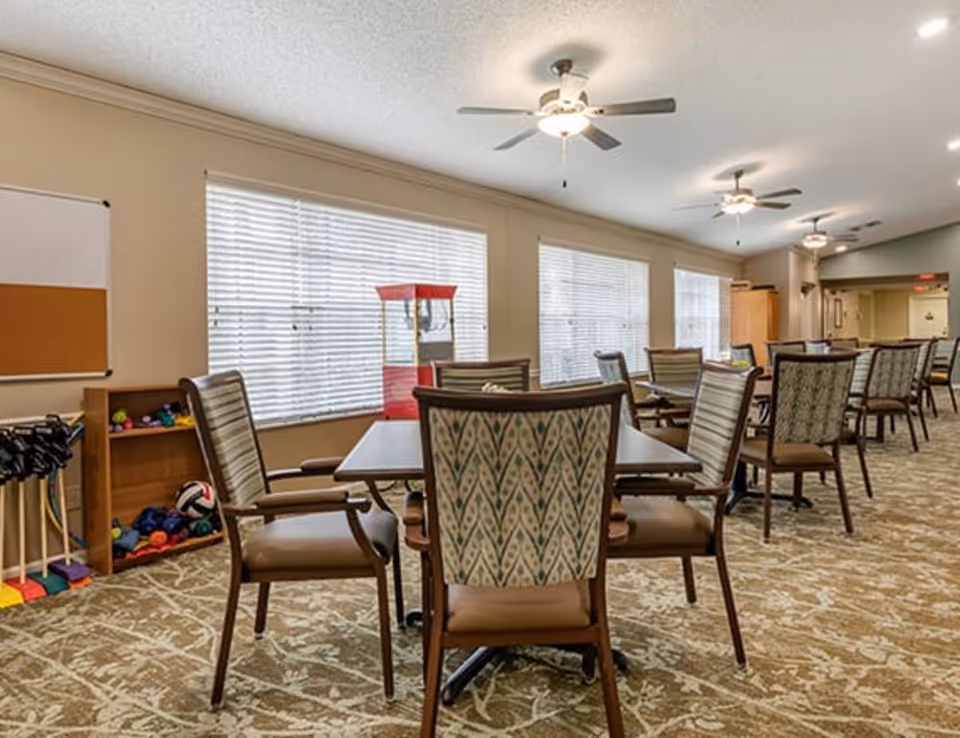 A spacious common area with several tables and chairs arranged in rows. The room has large windows with blinds, ceiling fans with lights, and a carpeted floor with a patterned design. In the corner, there is a small shelf with colorful balls and a red popcorn machine.