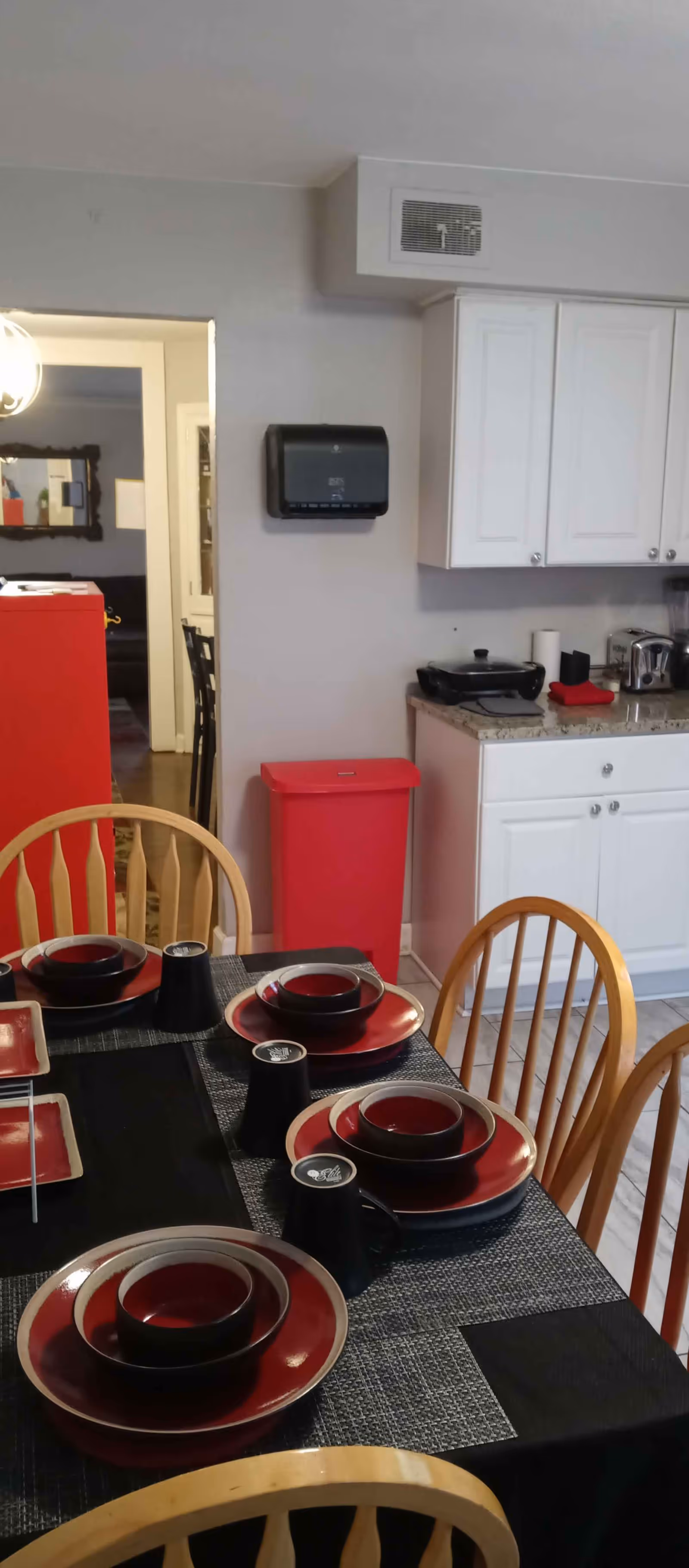 A dining area with a table set for four people with red and black plates, bowls, and mugs. The table has a black tablecloth and gray placemats. Behind the table is a kitchen area with white cabinets, a countertop with appliances, and a red trash bin. A doorway leads to another room with a mirror on the wall.