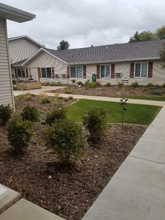 Outdoor view of a senior living facility with a landscaped garden area featuring small bushes and mulch beds, a concrete walkway, and a building with beige siding, maroon shutters, and several windows with air conditioning units.