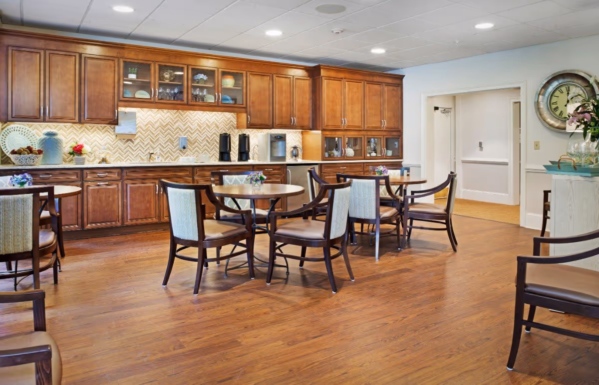 A dining area in a senior living facility with wooden floors and round tables surrounded by chairs. The back wall features wooden cabinets with glass doors, a countertop with a sink, coffee machines, and decorative items. A large clock and a flower arrangement are visible on the right side near an open doorway.