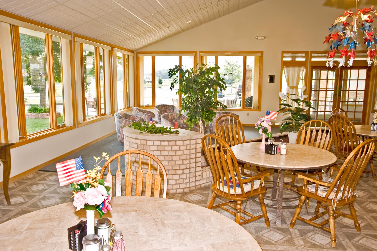 A bright dining area in a senior living facility with round tables and wooden chairs. Each table has a small floral centerpiece with an American flag. Large windows line one side of the room, allowing natural light to fill the space. There is a small brick planter with green plants and a seating area with cushioned chairs in the background. The ceiling is vaulted and decorated with red, white, and blue star-shaped hanging decorations.