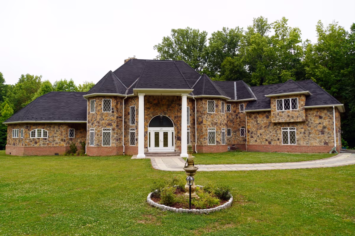 Front exterior view of a large stone house with multiple windows, white columns at the entrance, and a circular driveway with a small garden and fountain in the foreground, surrounded by green grass and trees.
