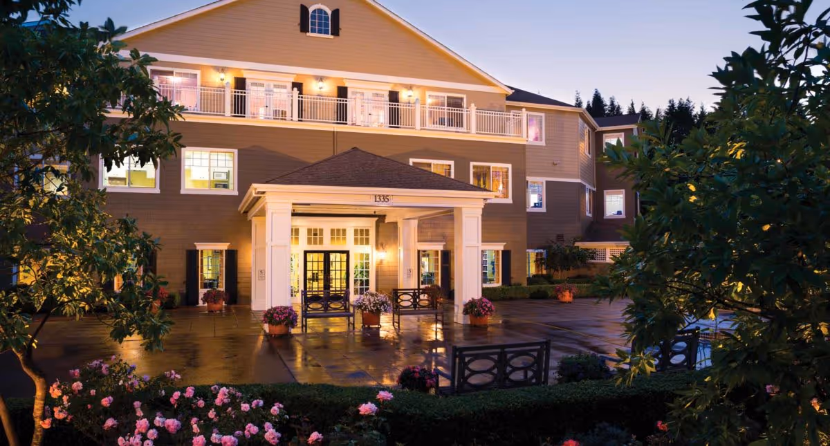 Exterior view of Canterbury Park Independent Living Community building at dusk, showing a three-story structure with lit windows, a covered entrance with benches, surrounded by trees and blooming flowers in the foreground.