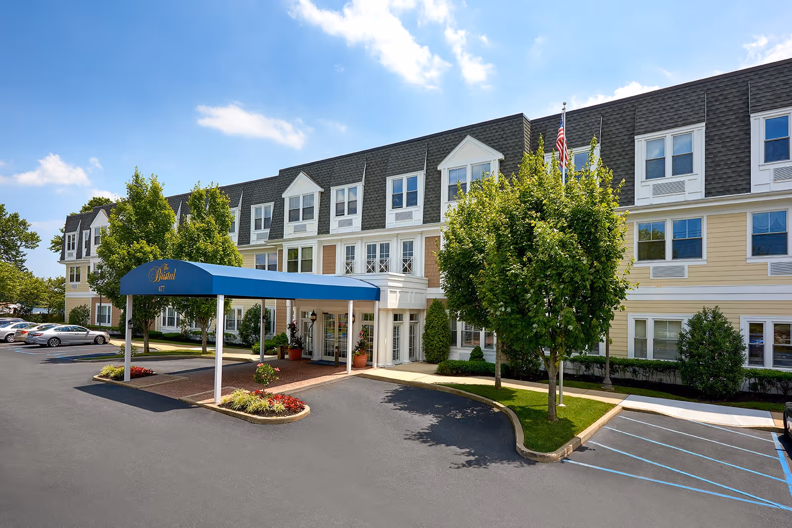 Exterior view of The Bristal Assisted Living at North Woodmere building with a blue entrance canopy, several trees, parked cars, and a clear blue sky.