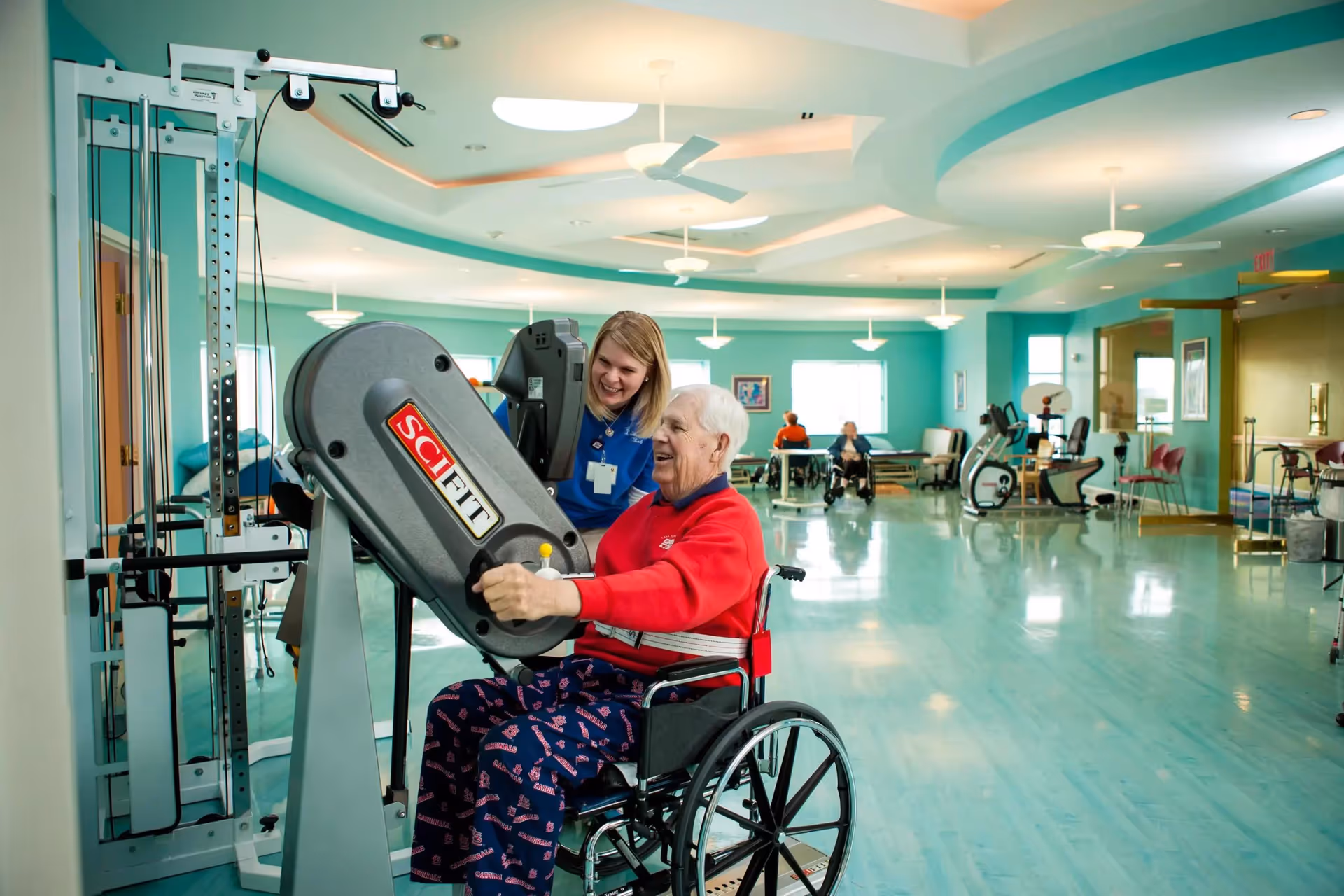 An elderly man in a wheelchair using a SCIFIT exercise machine in a spacious, brightly lit rehabilitation or fitness room with teal walls and ceiling fans. A female staff member in a blue uniform is smiling and assisting him. In the background, other elderly individuals are seated and engaged in activities.