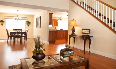 A bright and spacious interior of a senior living facility showing a wooden dining table with four chairs near a large window, a kitchen area with wooden cabinets in the background, and a living room area with a glass-top coffee table adorned with a decorative plant and a small ornate box. A wooden staircase with white balusters is visible on the right side, along with a small side table holding a lamp and a framed sign.