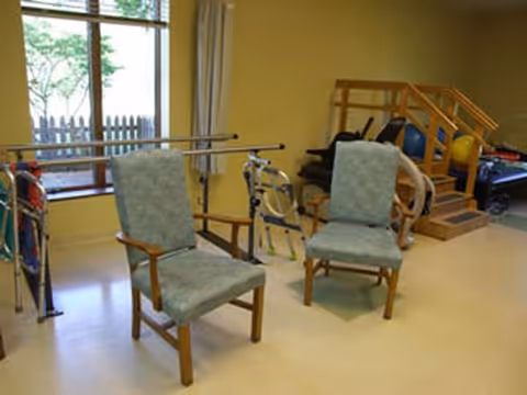 Two cushioned chairs in an interior rehab room with parallel bars and equipment.