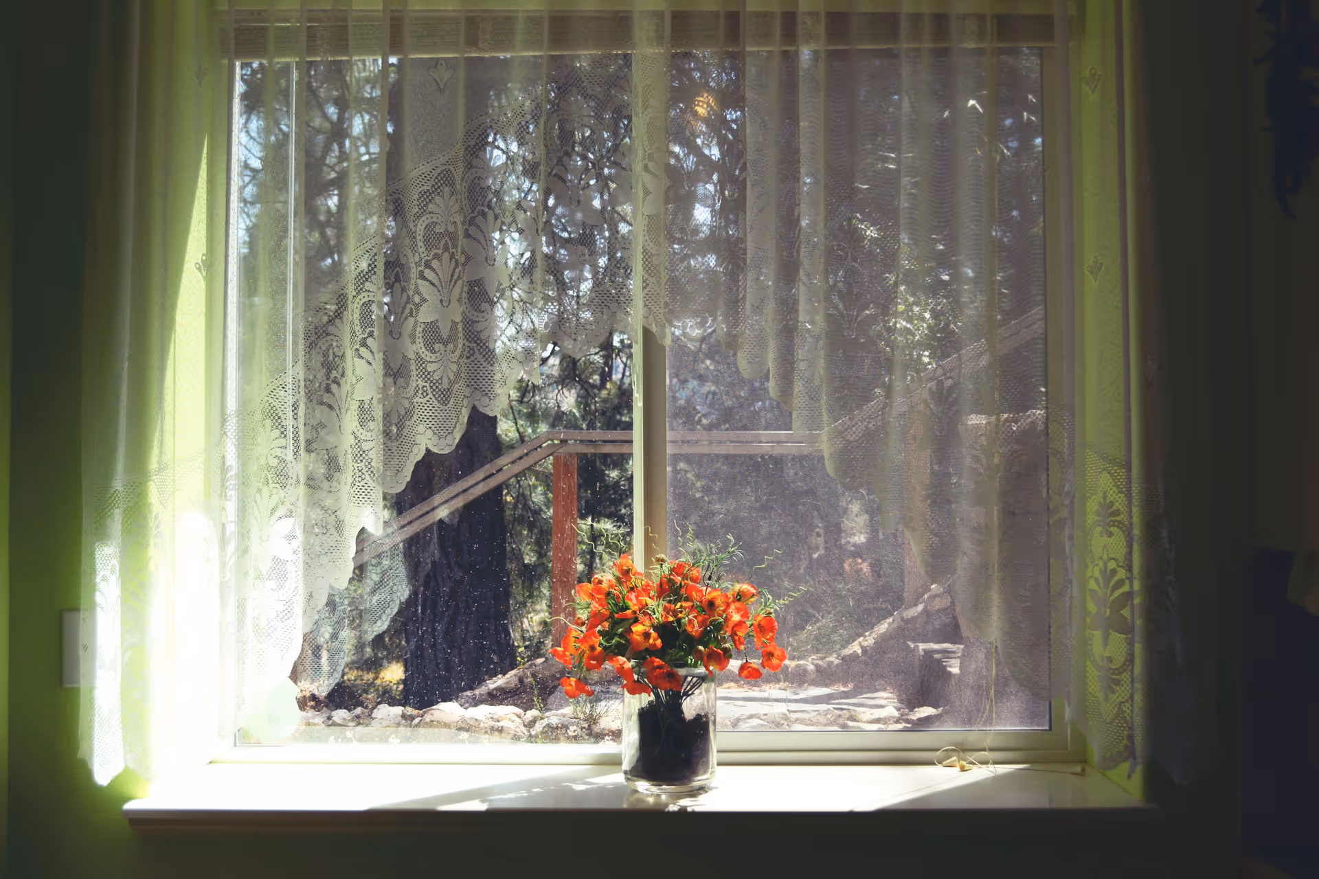 A window with sheer lace curtains partially drawn to the sides, allowing sunlight to filter through. On the windowsill, there is a glass vase filled with vibrant orange flowers. Outside the window, trees and a wooden railing are visible.