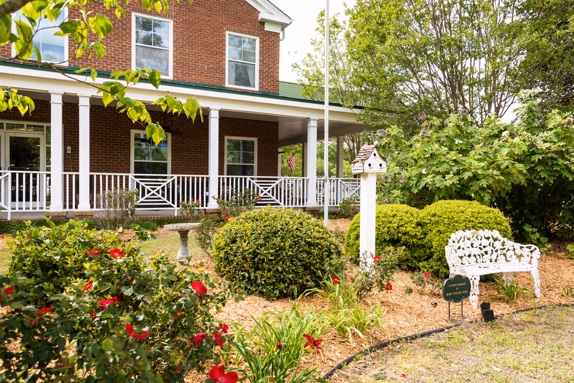 Brick building front with a covered porch and white railing, surrounded by a landscaped garden with shrubs, red flowers, a birdhouse and a white bench.