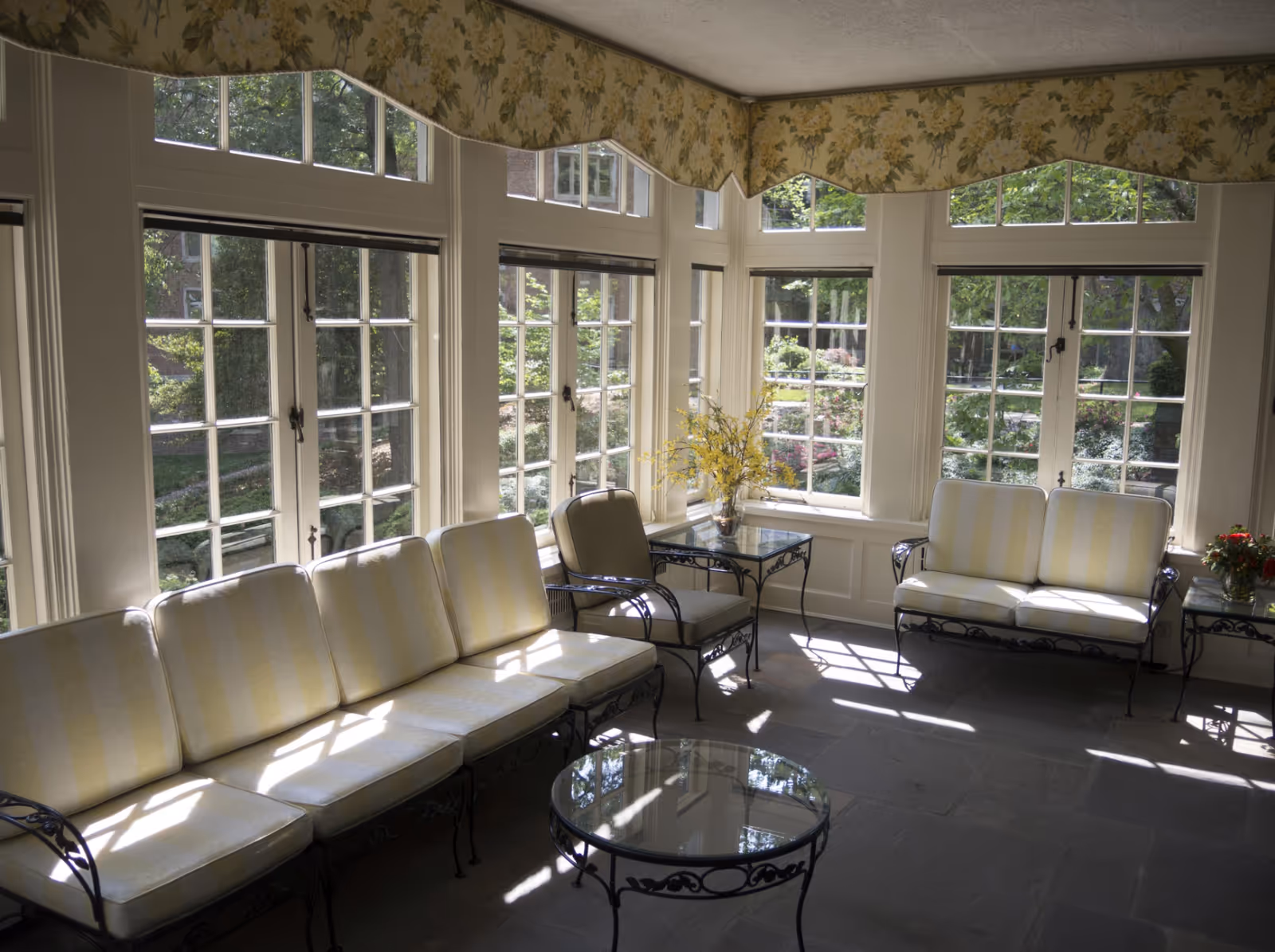 Sunlit sitting area with cushioned wrought iron furniture including a long sofa, a loveseat, and a single chair arranged around glass-top tables. The room features large windows with white frames and floral valances, offering a view of a garden outside.