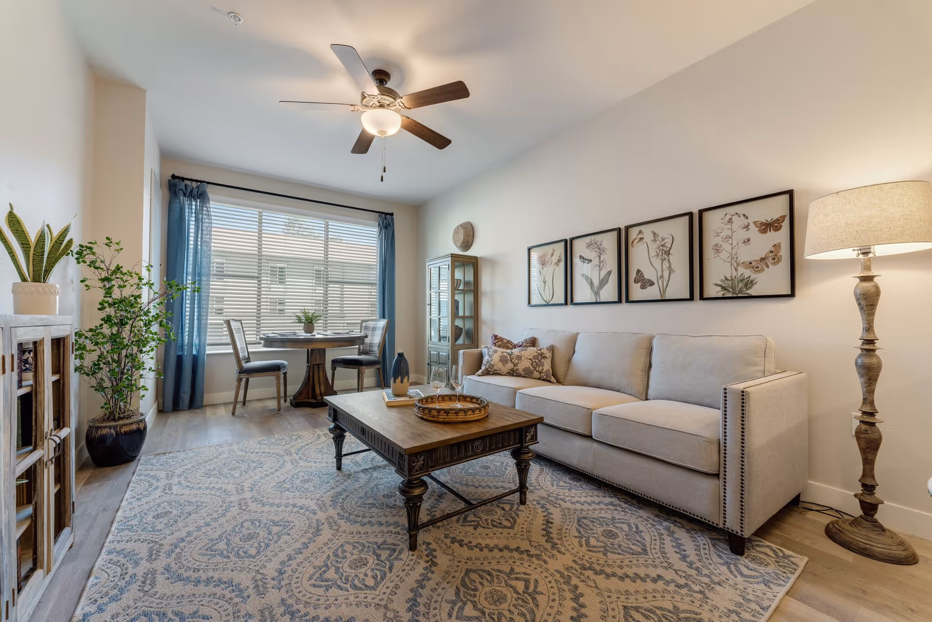 A cozy senior living room featuring a beige sofa with decorative pillows, a wooden coffee table with a tray and glasses, a patterned area rug, a tall floor lamp, and framed botanical artwork on the wall. In the background, there is a round dining table with two chairs near a large window with blue curtains, and a ceiling fan with a light fixture above.