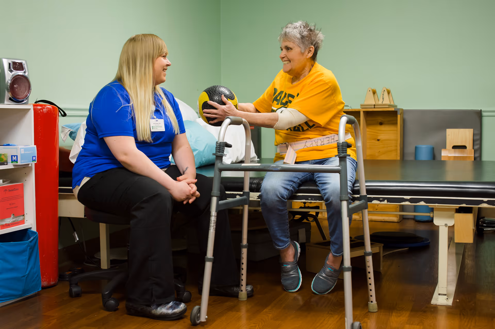A senior woman sitting on a therapy table holding a medicine ball and smiling at a female caregiver or therapist who is seated nearby in a room with light green walls and wooden flooring.