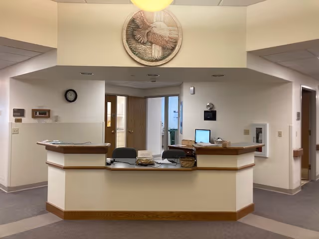 Reception desk area inside a senior living facility with two chairs behind the desk, a computer monitor, a basket, and a round decorative wall art featuring a bird above the desk. Doors and hallway are visible in the background.
