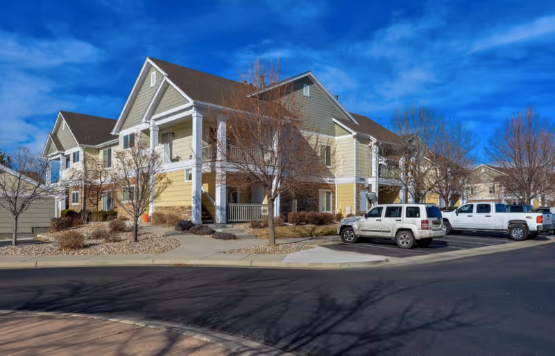 Exterior view of a two-story residential building with beige and yellow siding, white trim, and a brown roof under a bright blue sky. There are leafless trees and landscaped areas with rocks and shrubs around the building. Several vehicles are parked in the parking lot in front of the building.