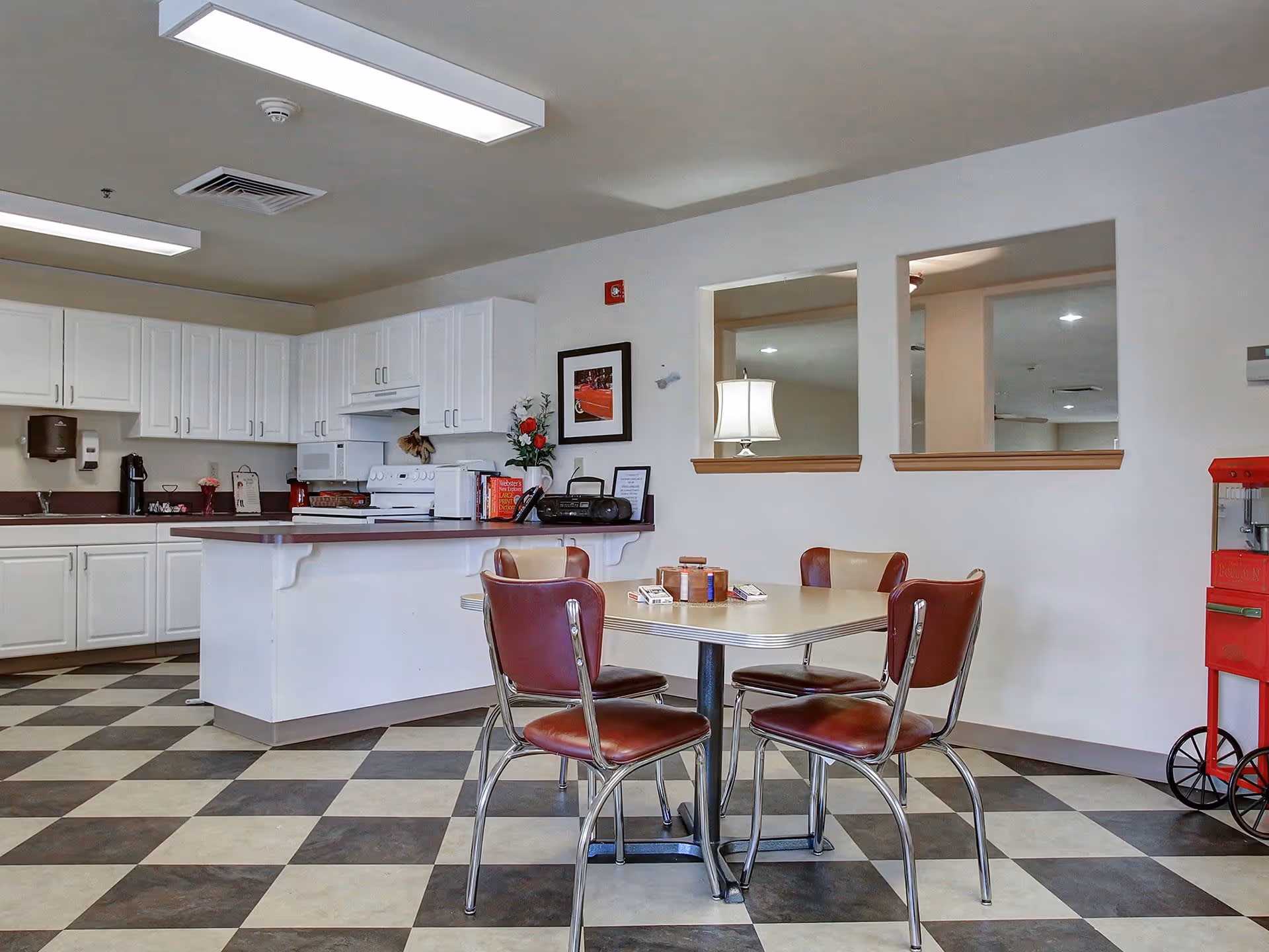 A senior living facility kitchen and dining area with white cabinets, a white stove, microwave, and countertop. There is a square dining table with four retro-style chairs with red and beige upholstery. The floor has a black and white checkered pattern. A red popcorn machine is visible on the right side, and two rectangular windows in the wall show a lamp and ceiling fan in the adjacent room.