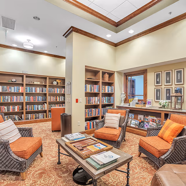 A cozy library room with bookshelves filled with books along the walls. There are four wicker armchairs with orange cushions arranged around a wooden coffee table that holds books and a backgammon game. The room has beige walls, a patterned carpet, and wood trim accents. Decorative framed art and a window with a counter displaying magazines and flowers are also visible.