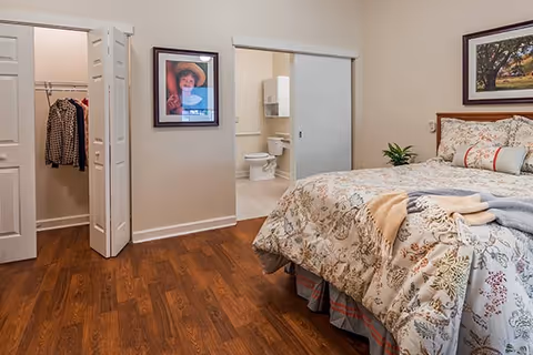 Well-lit bedroom with a patterned bed, hardwood floors, open closet doors and an adjoining bathroom visible through a sliding door.