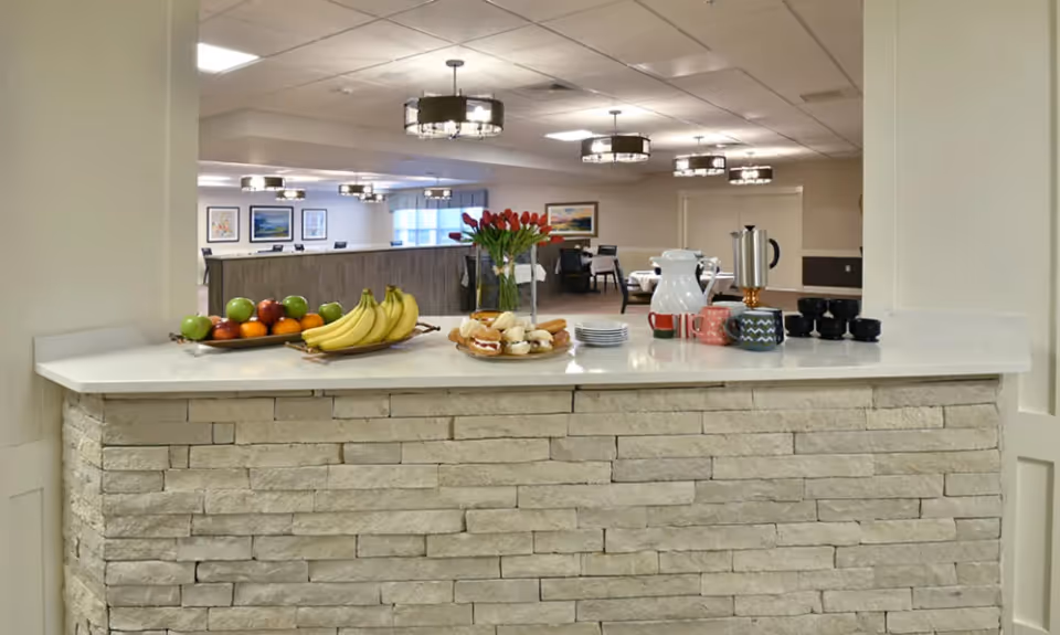 A serving counter with a stone facade topped with a white countertop displaying a tray of assorted fruits including bananas, apples, and oranges, a plate of pastries, a vase with red tulips, a white pitcher, a coffee urn, colorful mugs, and black cups. In the background, a dining area with tables, chairs, framed artwork on the walls, and ceiling lights is visible.