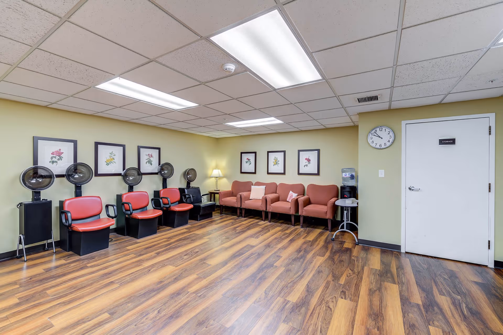 Interior room with wooden flooring and light green walls featuring four red salon hair dryer chairs on the left side and four pink cushioned armchairs along the back wall. The walls are decorated with framed floral artwork, and there is a standing lamp in the corner. A white door labeled 'STORAGE' and a wall clock are visible on the right side, along with a water dispenser and a small round table.