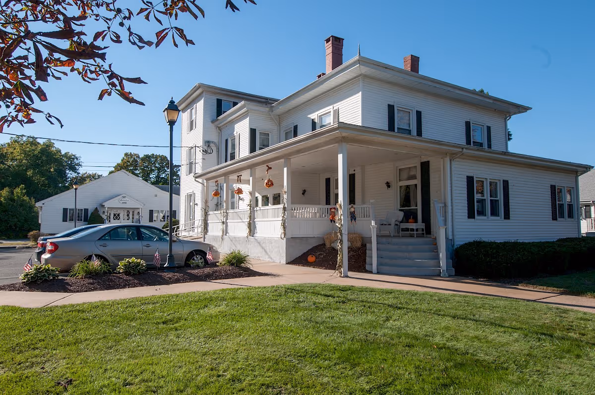 Exterior view of a large white two-story house with a wraparound porch decorated with autumn-themed hanging decorations and pumpkins. There is a car parked near the porch, a streetlamp, and a well-maintained lawn in the foreground under a clear blue sky.