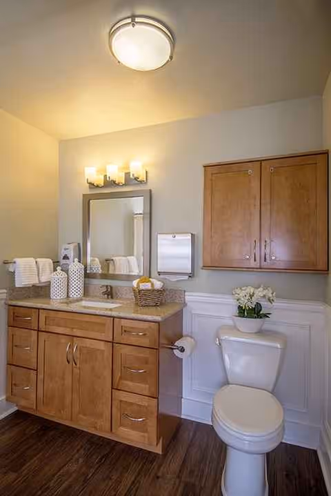 Well-lit bathroom with wooden vanity, mirror, towels, and a toilet.