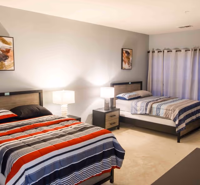 A bedroom with two beds, each with striped bedding in different color schemes. There are two nightstands between the beds, each with a lamp. The room has light-colored walls, two framed abstract paintings above the beds, and a window with closed curtains.