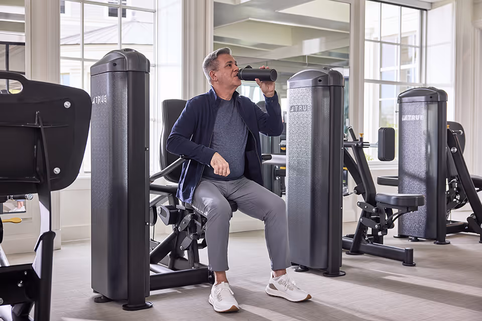A man seated on exercise equipment in a bright fitness room drinking from a water bottle.