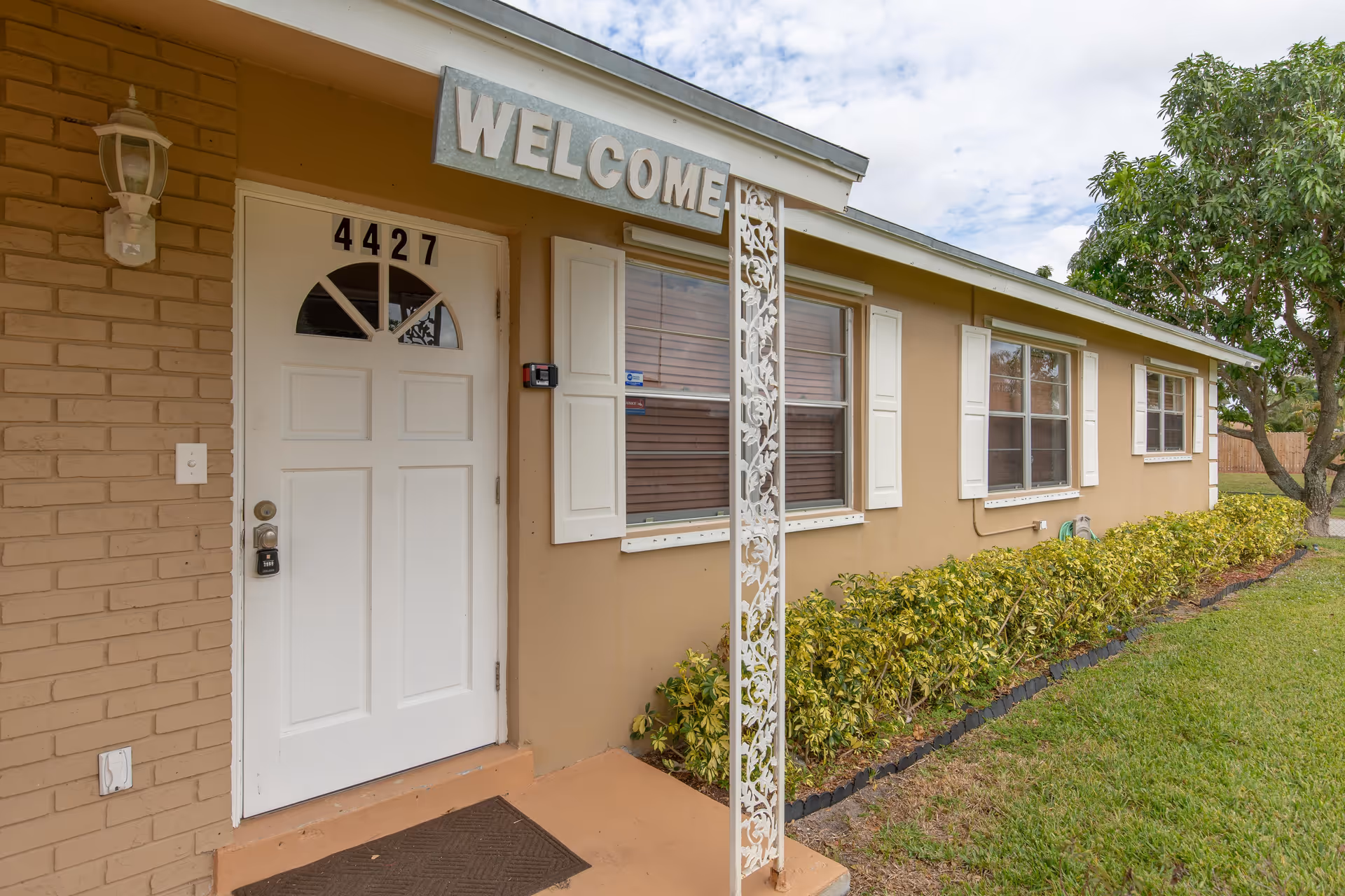 Exterior view of a single-story building with a white door numbered 4427, a 'WELCOME' sign above the door, beige walls, white window shutters, a decorative white metal post, and a small garden with green shrubs and a tree on the right side.