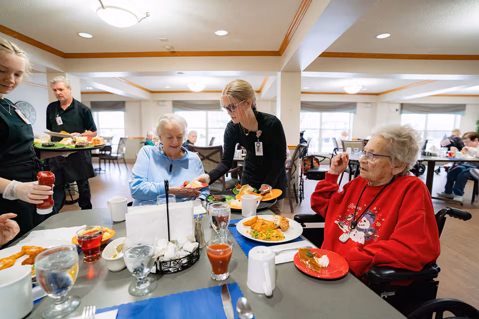Two elderly women seated at a dining table in a senior living facility, being served food by two staff members. The table has plates with meals, glasses of water, and condiments. Other residents and staff are visible in the background in a bright dining room with large windows.