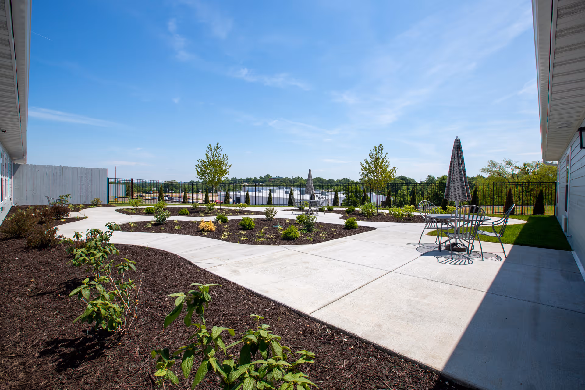 Outdoor patio area at Oak Pointe of Washington featuring concrete walkways, planted garden beds with small shrubs and trees, metal tables with chairs, and closed umbrellas under a clear blue sky.