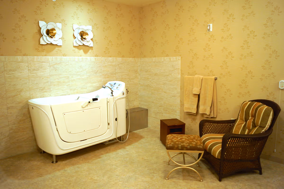 A bathroom with a walk-in bathtub, beige tiled walls, and floral wallpaper above the tiles. There are two decorative white and gold flower wall hangings above the bathtub. To the right, there is a cushioned wicker chair with a striped cushion and a matching footstool. Two beige towels hang on a towel rack on the wall.