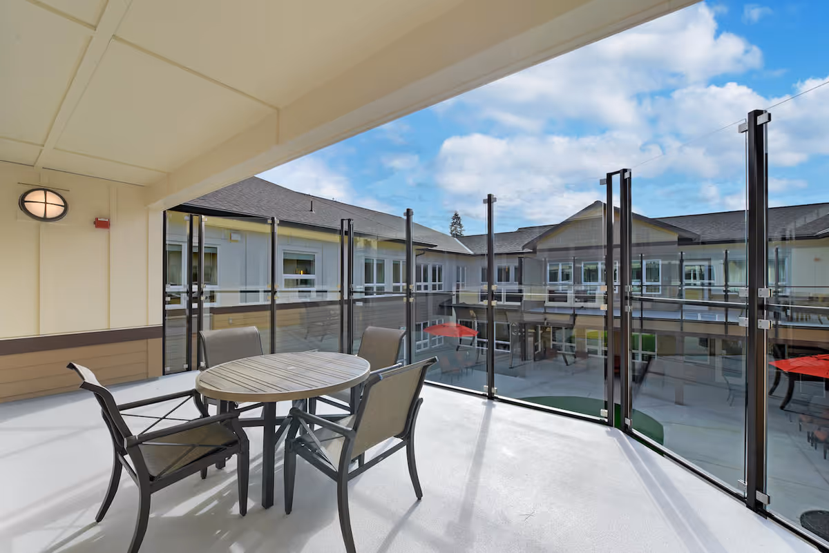 Covered outdoor balcony area with a round table and four chairs, enclosed by glass panels. The balcony overlooks a courtyard of a multi-story building under a partly cloudy sky.