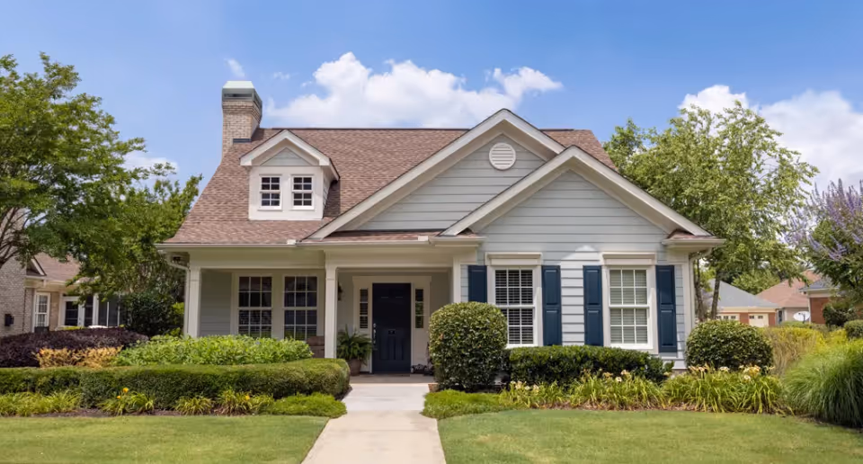 Front exterior view of a single-story house with gray siding, a brown shingled roof, white trim, and blue shutters. The house has a small covered porch with a black front door, surrounded by well-maintained green shrubs and a manicured lawn. Trees and neighboring houses are visible in the background under a partly cloudy sky.
