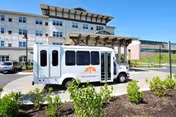 A white shuttle bus with the Covenant Village logo parked in front of a large multi-story senior living facility building with a covered entrance and landscaped greenery in the foreground.