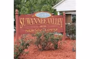A red wooden sign with gold lettering that reads 'Suwannee Valley Nursing Center' placed in a landscaped area with mulch and bushes, with part of a building and trees visible in the background.