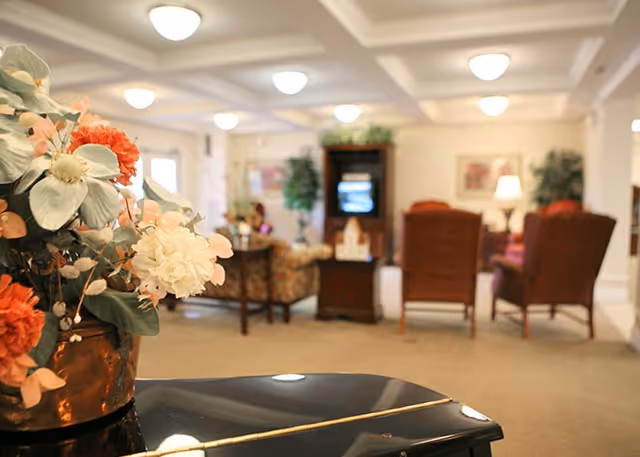 Floral arrangement on a piano in the foreground with a softly lit lounge area, armchairs, and a TV in the background.