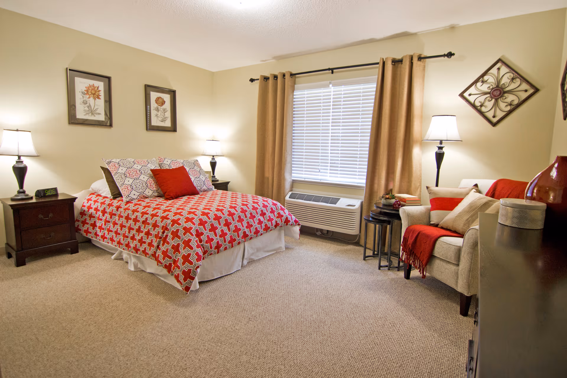 A cozy bedroom with a bed covered in a red and white patterned quilt, two bedside tables each with a lamp, a window with beige curtains, an air conditioning unit below the window, a beige armchair with red and beige cushions and a red throw, and decorative wall art.