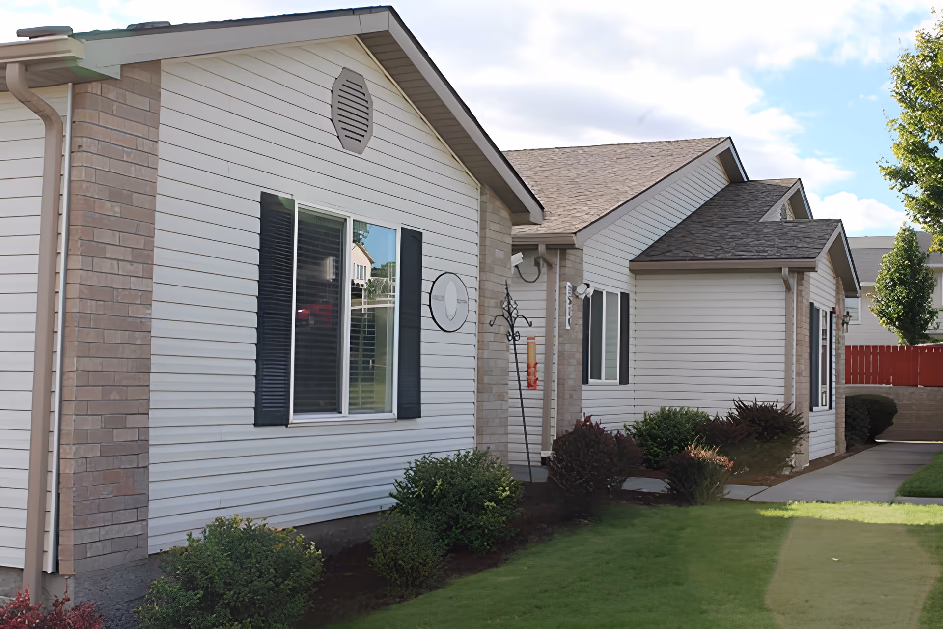 Exterior view of a single-story building with white siding and brick accents, featuring windows with black shutters, a well-maintained lawn, shrubs, and a concrete walkway under a partly cloudy sky.