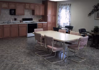 A dining area in an assisted living facility featuring a rectangular table with eight chairs around it. The room has a kitchen area with wooden cabinets, a stove, and a sink. There is a window with floral curtains and a small table with a plant and decorative items on the right side.