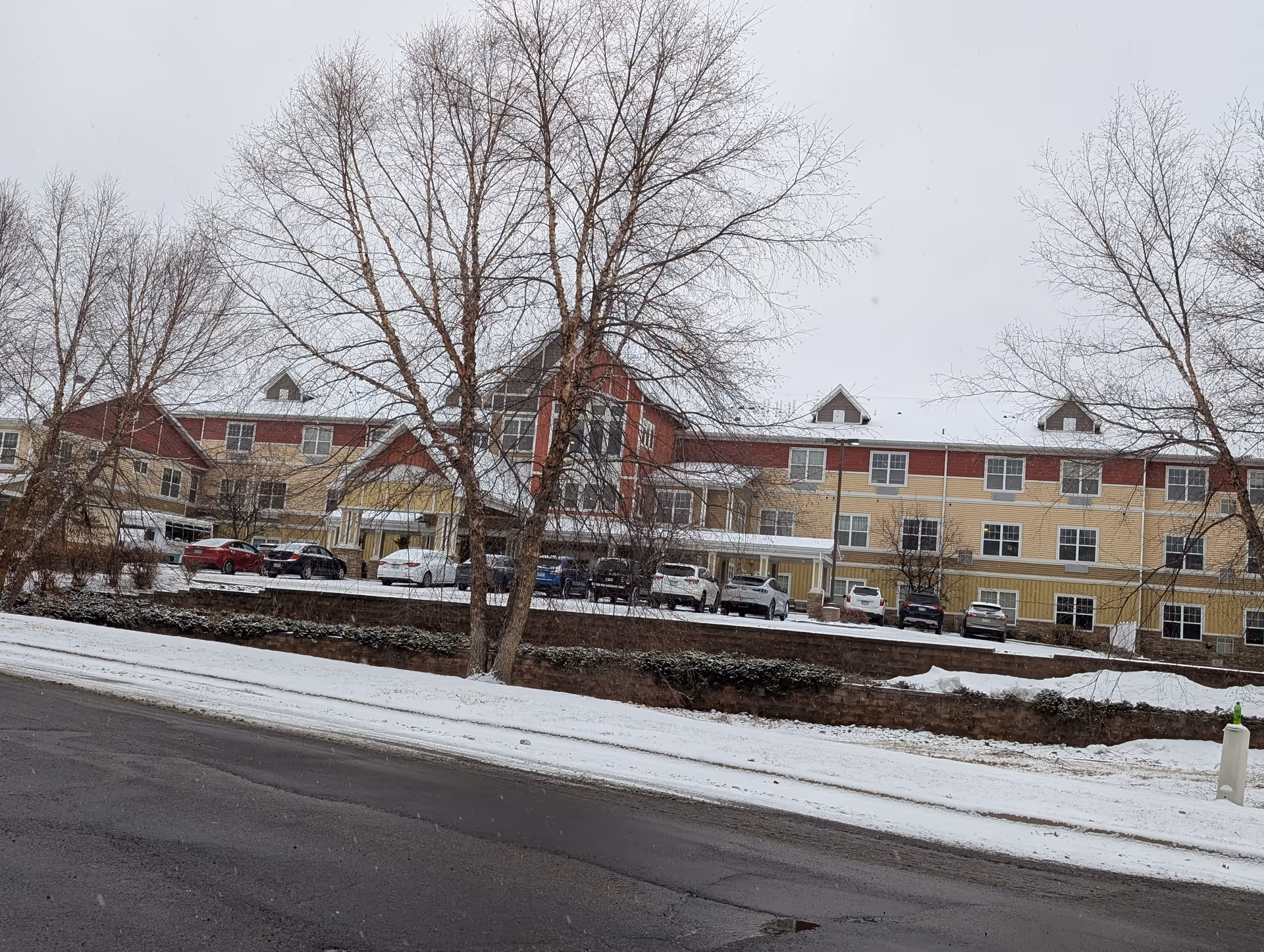 Exterior view of a senior living facility building with a red and yellow facade, multiple windows, and a covered entrance. Several cars are parked in front of the building. The ground and trees are covered with a light layer of snow, and the sky is overcast.