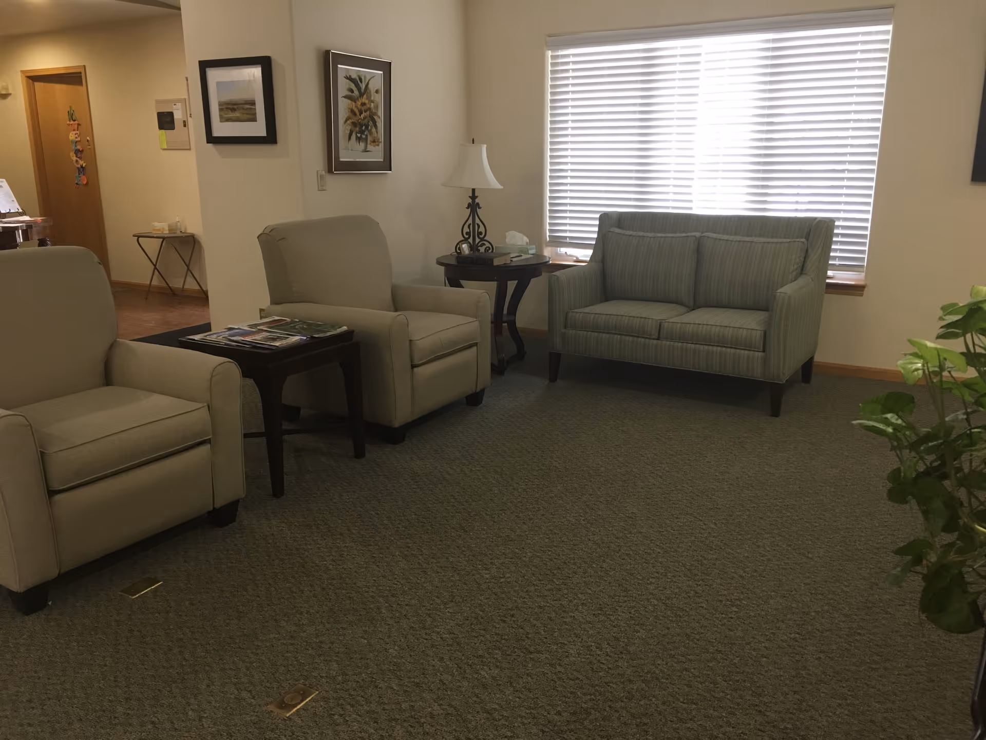 A cozy sitting area in a senior living facility with two beige armchairs and a green striped loveseat arranged around two small wooden side tables. One table has a lamp and tissues, and the other has magazines. The room has carpeted flooring, a large window with blinds, framed artwork on the walls, and a plant partially visible in the foreground.