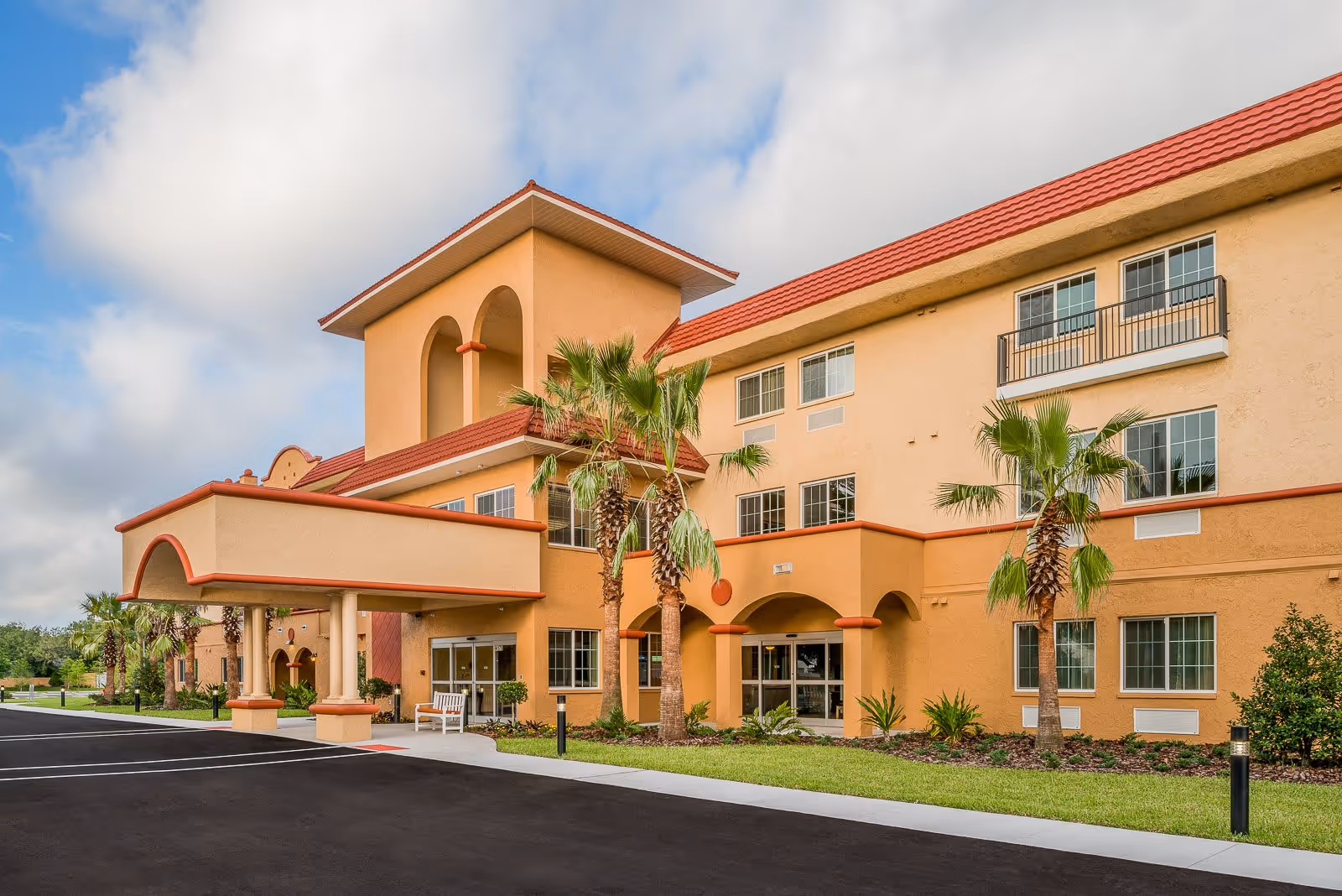 Exterior view of a three-story senior living facility building with a covered entrance, palm trees, and a well-maintained lawn under a partly cloudy sky.