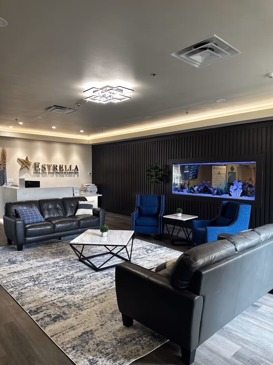 A modern and comfortable lounge area in Estrella Health and Rehab Center featuring two black leather sofas, two blue upholstered armchairs, a marble coffee table, and a matching side table. The room has a large area rug with a gray and white pattern, a dark paneled accent wall with a built-in aquarium, and a reception desk with the Estrella Health and Rehab Center logo on the wall behind it. The ceiling has recessed lighting and a contemporary square light fixture.