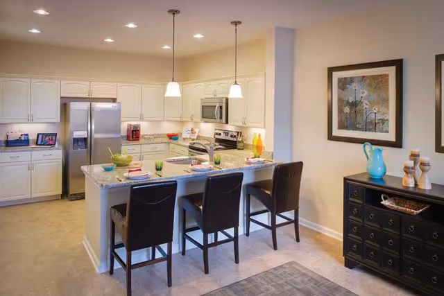 A modern kitchen with white cabinets, stainless steel refrigerator, microwave, and stove. A granite countertop island with three dark brown chairs is set with plates and glasses. Pendant lights hang above the island. A black sideboard with decorative items and a framed floral painting are on the adjacent wall.