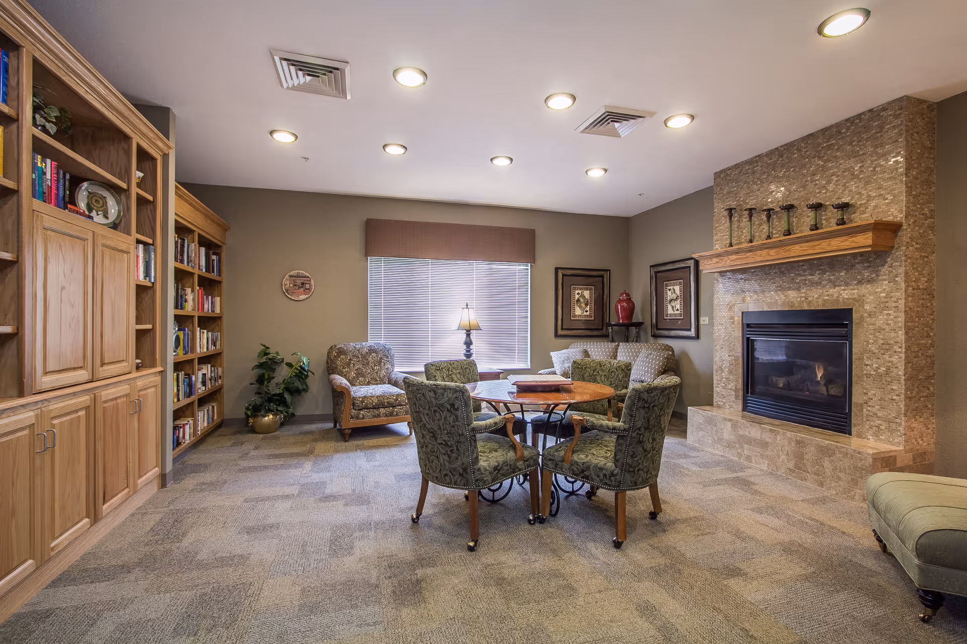 A cozy senior living common area with a round wooden table surrounded by four upholstered chairs, a fireplace with a tiled surround and wooden mantel, a bookshelf filled with books, two armchairs, a sofa, framed artwork on the walls, a window with blinds and a lamp on a side table.